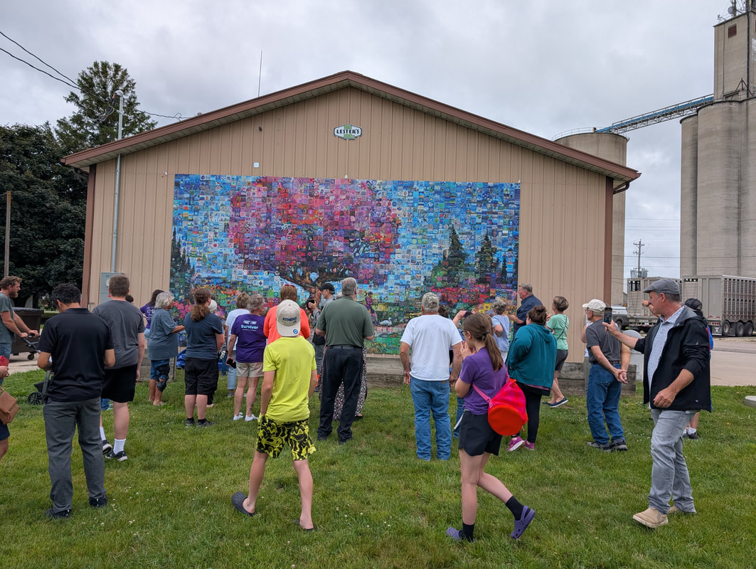Crowd stands on grass facing a large barn wall covered with a colorful photo mosaic mural.